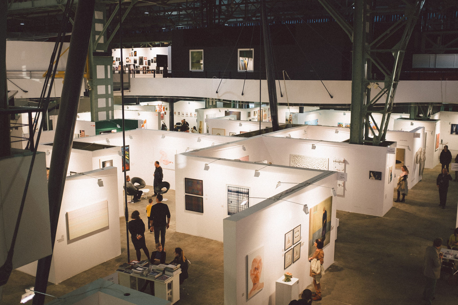 Visitors browsing artwork in exhibition booths inside a large industrial hall.