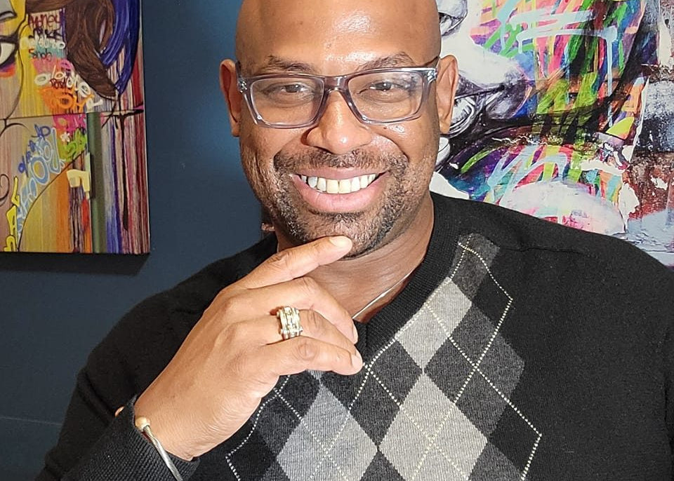 Corey Wesley wearing glasses and a black sweater with a checkered pattern, standing in front of colorful artwork.