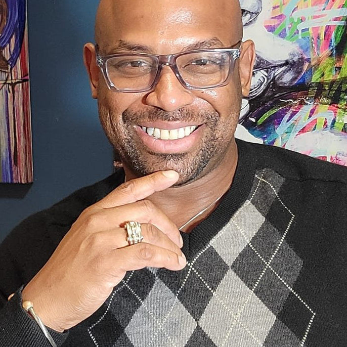 Corey Wesley wearing glasses and a black sweater with a checkered pattern, standing in front of colorful artwork.