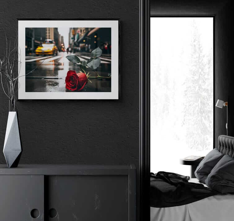 Framed poster of a red rose on a rainy NYC street, displayed on a dark wall in a modern bedroom, promoting Afrocentric home decor and colorful wall art.