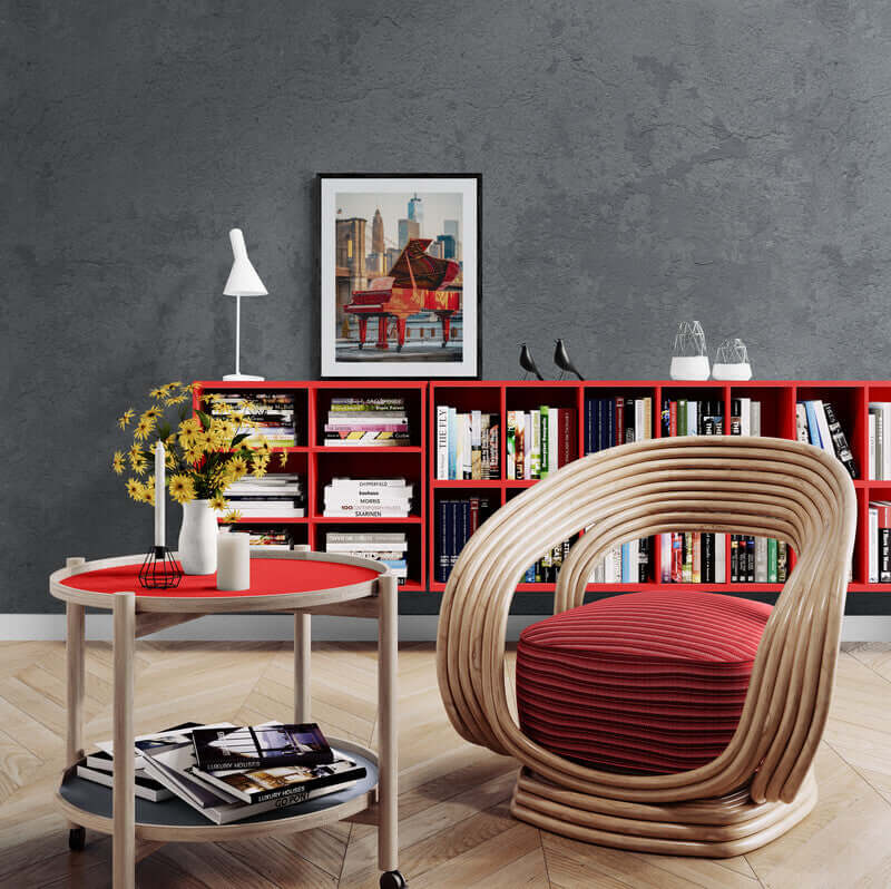 Modern living room with red and wood accents, featuring a framed cityscape print, bookshelf, and rattan chair.