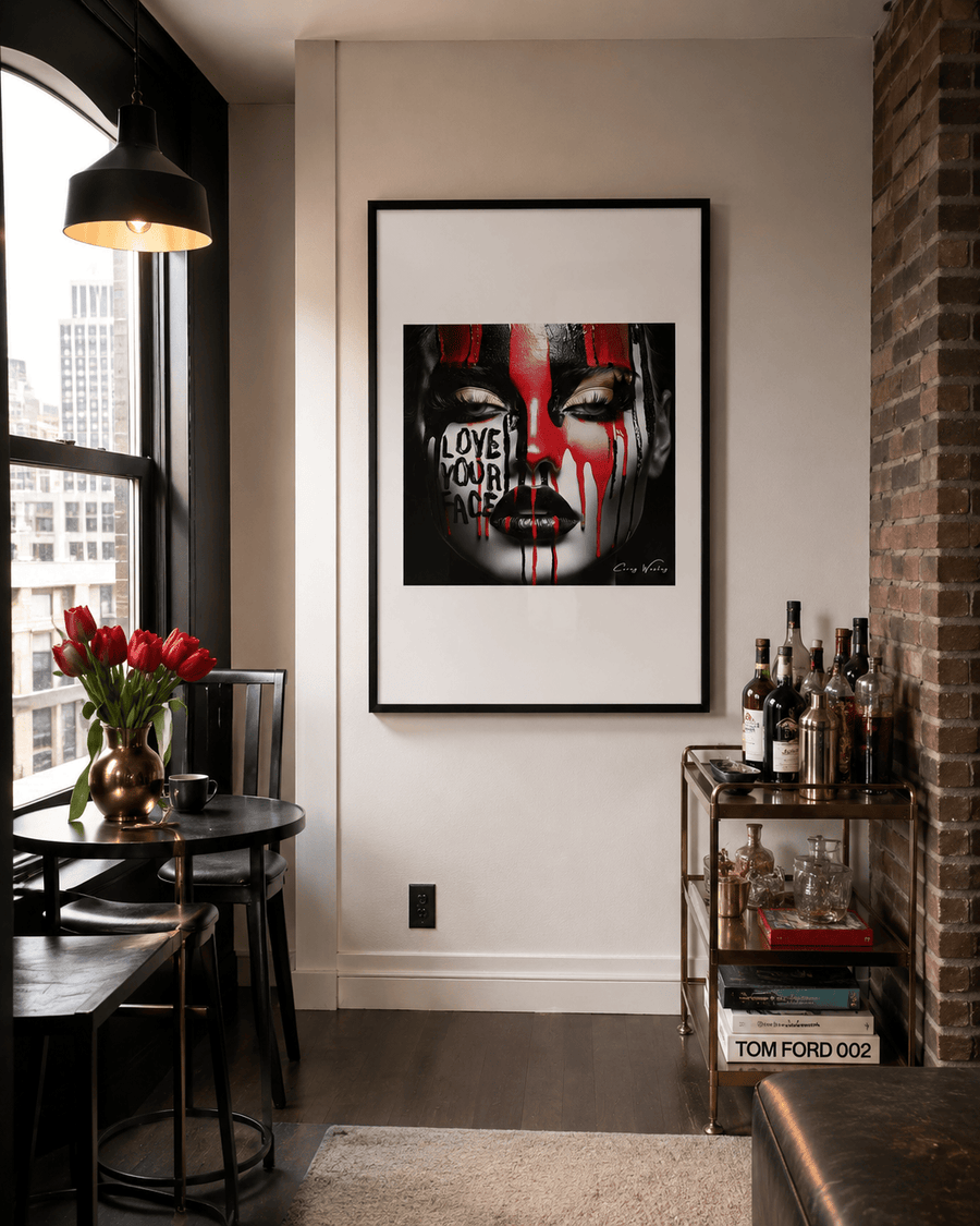 A framed one-of-one piece from Milton Wes Art's Unapologetic Faces series hangs on a light gray wall in a Harlem apartment dining nook, above a bar cart. The archival museum-quality aluminum work depicts a close-up face in black and white with bold red and black paint streaks running down the forehead and cheeks, dripping over closed eyes and lips, with the words "LOVE YOUR FACE" written across the mouth. The piece is displayed in a black frame at commanding scale near a window with a city skyline view